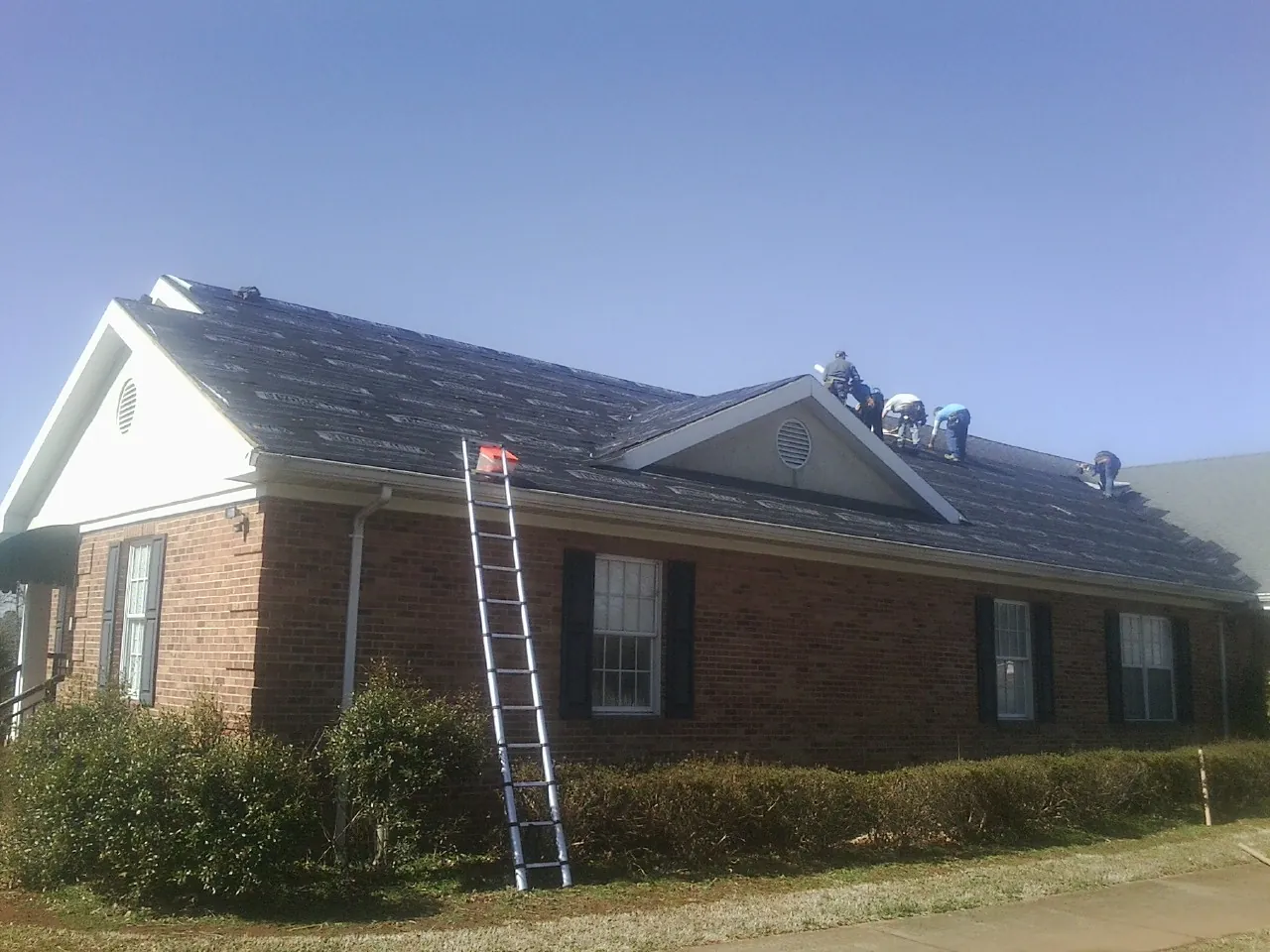 Crew tearing shingles off a brick ranch roof with a ladder set — Emergency Roof Repair work by our crew in Hampstead