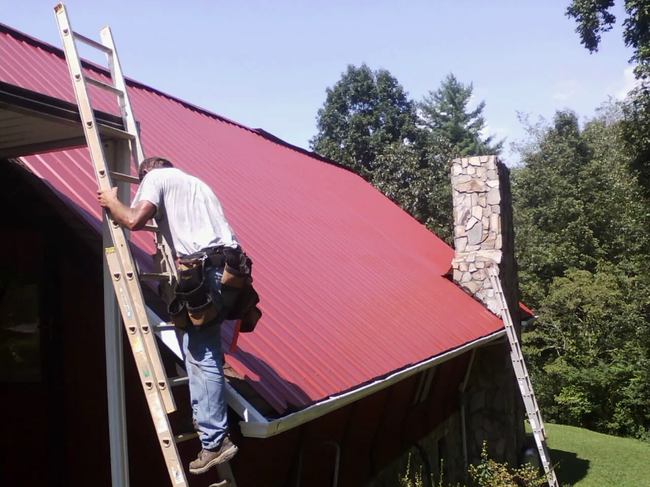 Roofer on a ladder beside a red metal roof and stone chimney — Soffit & Fascia Repair by Roofing Brothers in Hampstead